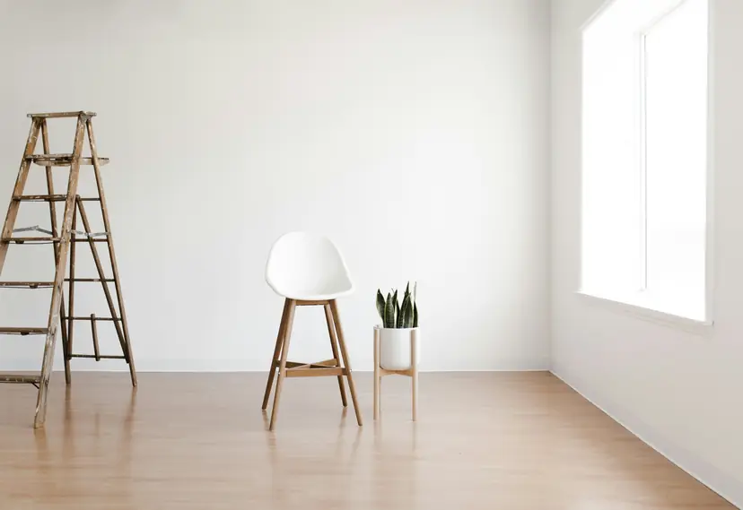 Habitación luminosa y vacía con una escalera de madera, un taburete blanco y una sansevieria junto a la ventana