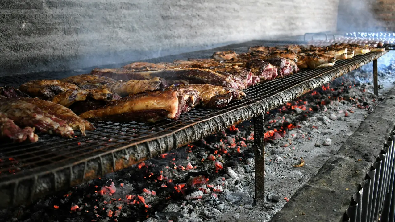 A long charcoal grill loaded with cuts over glowing embers, smoke against a brick wall